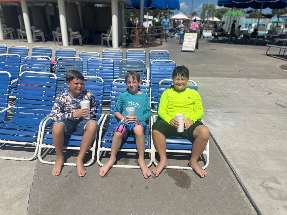 Three boys in swim shirts sit on blue lounge chairs at a water park, holding large drinks